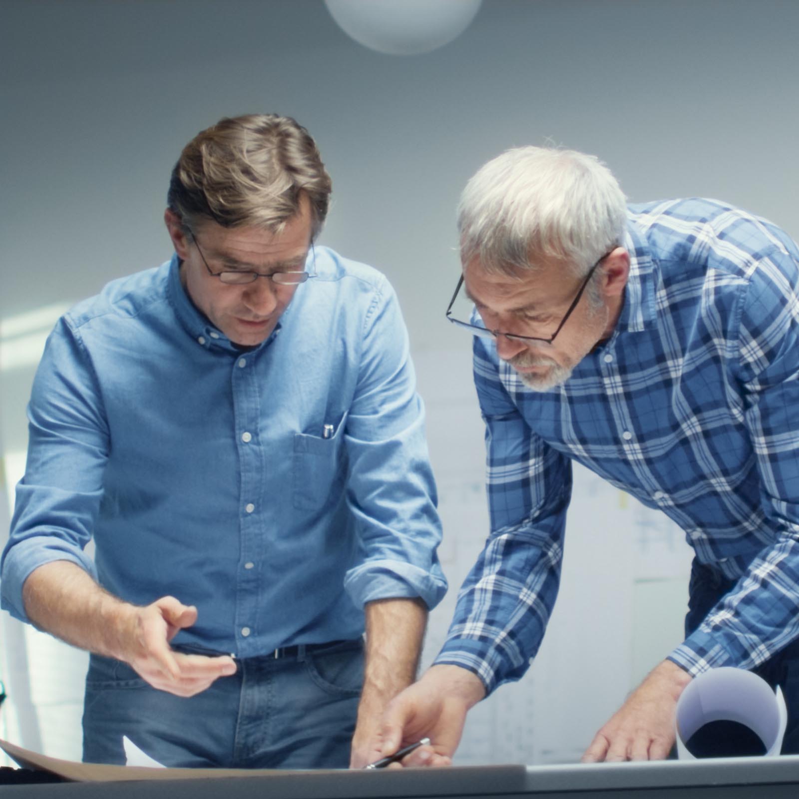 Two Senior Engineers Work with Schemes and Blueprints while Standing over Working Table. They have Discussion on the Subject of Various Technical Matters Concerning Their Project.